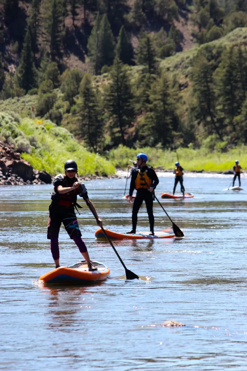 Rafting safety gear including life jackets, helmets, paddles, and dry bags arranged on a dock near the Colorado River